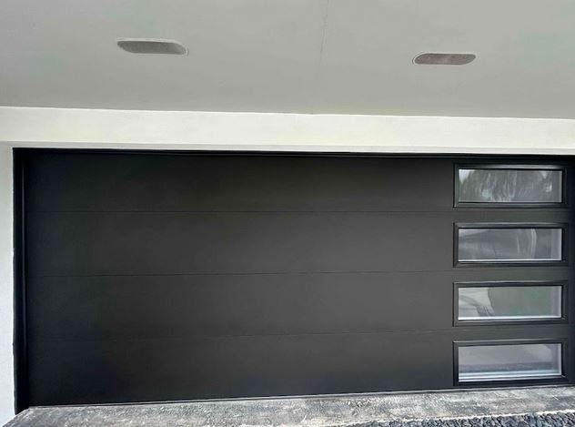 This black garage door features a modern steel design with a row of slim, horizontal windows on the right side.